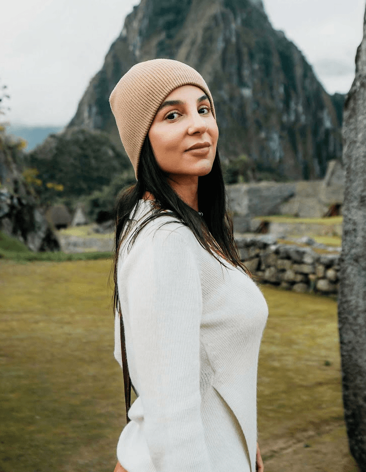 Smiling young woman with long hair standing against a dark green background, holding a finger to her chin.