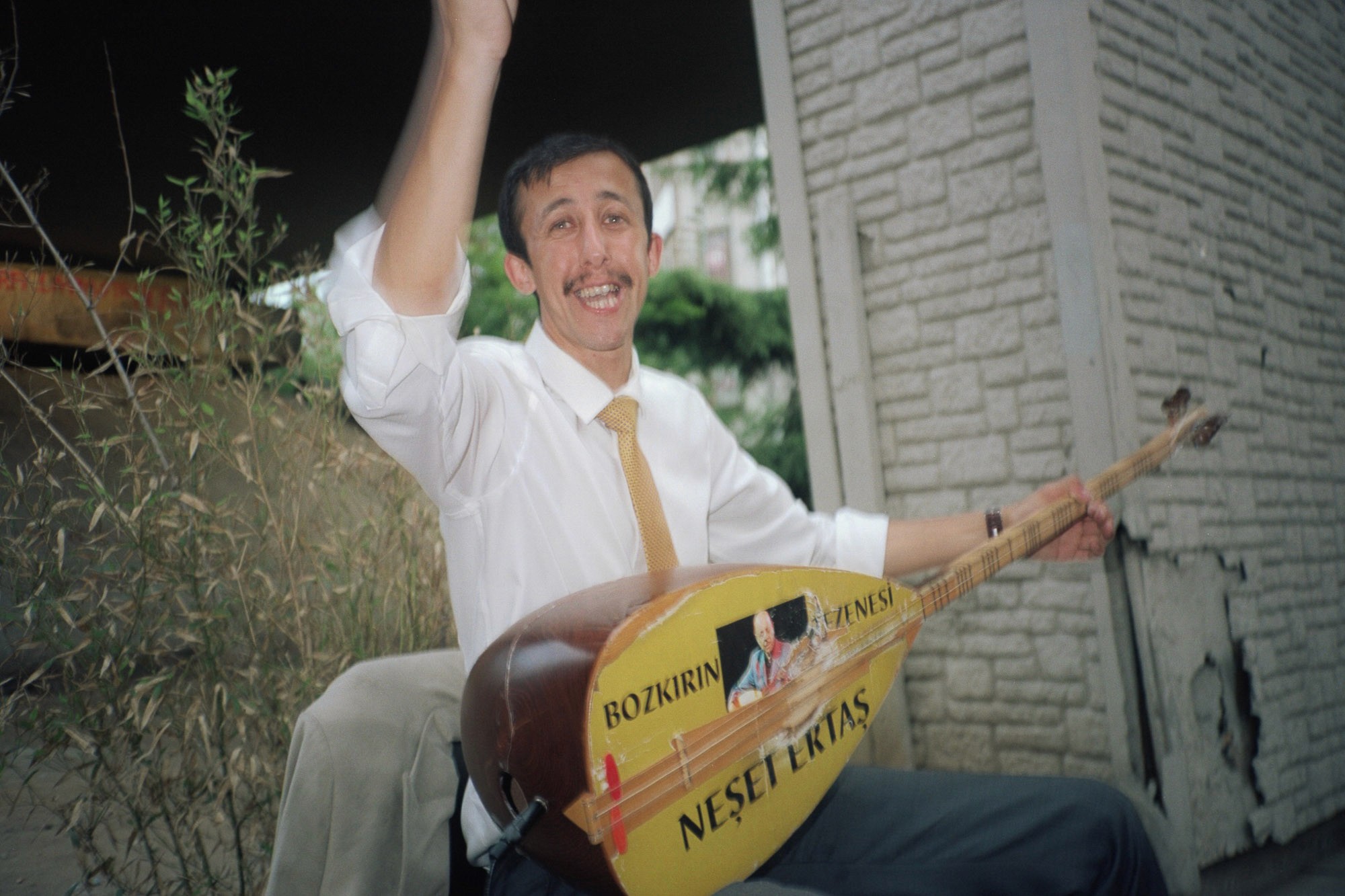 A joyful man sitting outdoors plays a traditional stringed instrument, marked with the name "Neşet Ertaş", against a backdrop of a stone wall and greenery.
