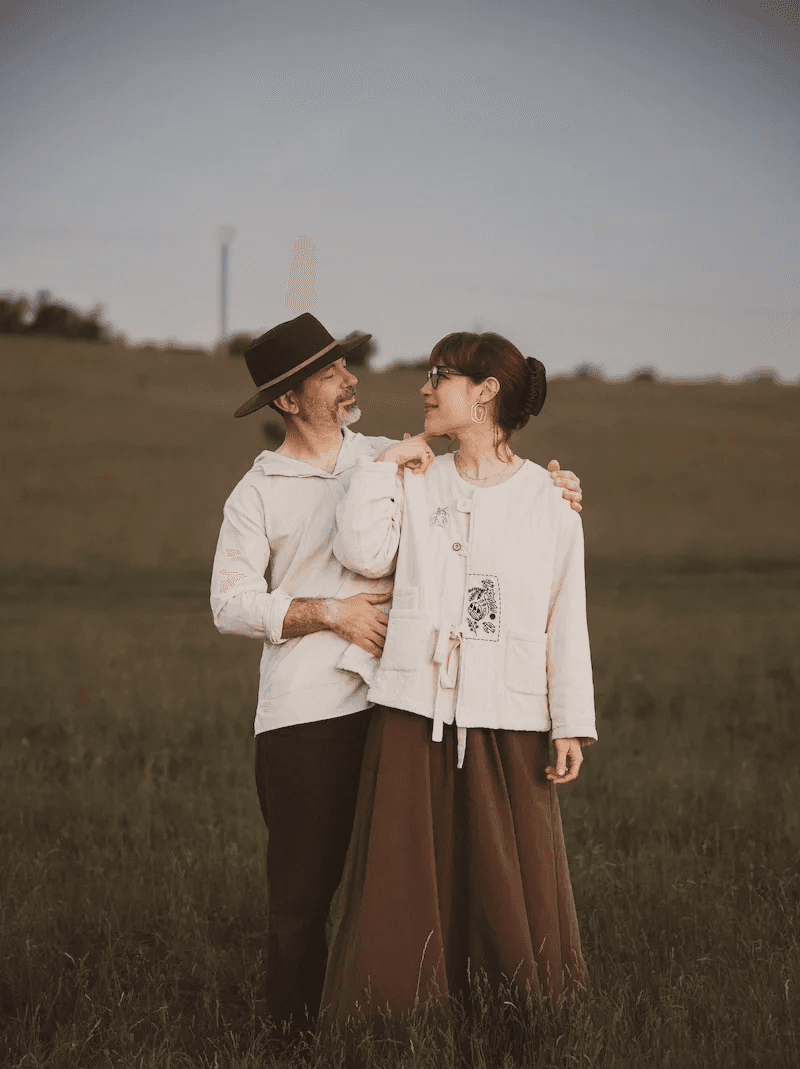 Cosmin și Ioana zâmbind unul la celălalt, îmbrăcați în haine luminoase pe un fundal de câmp