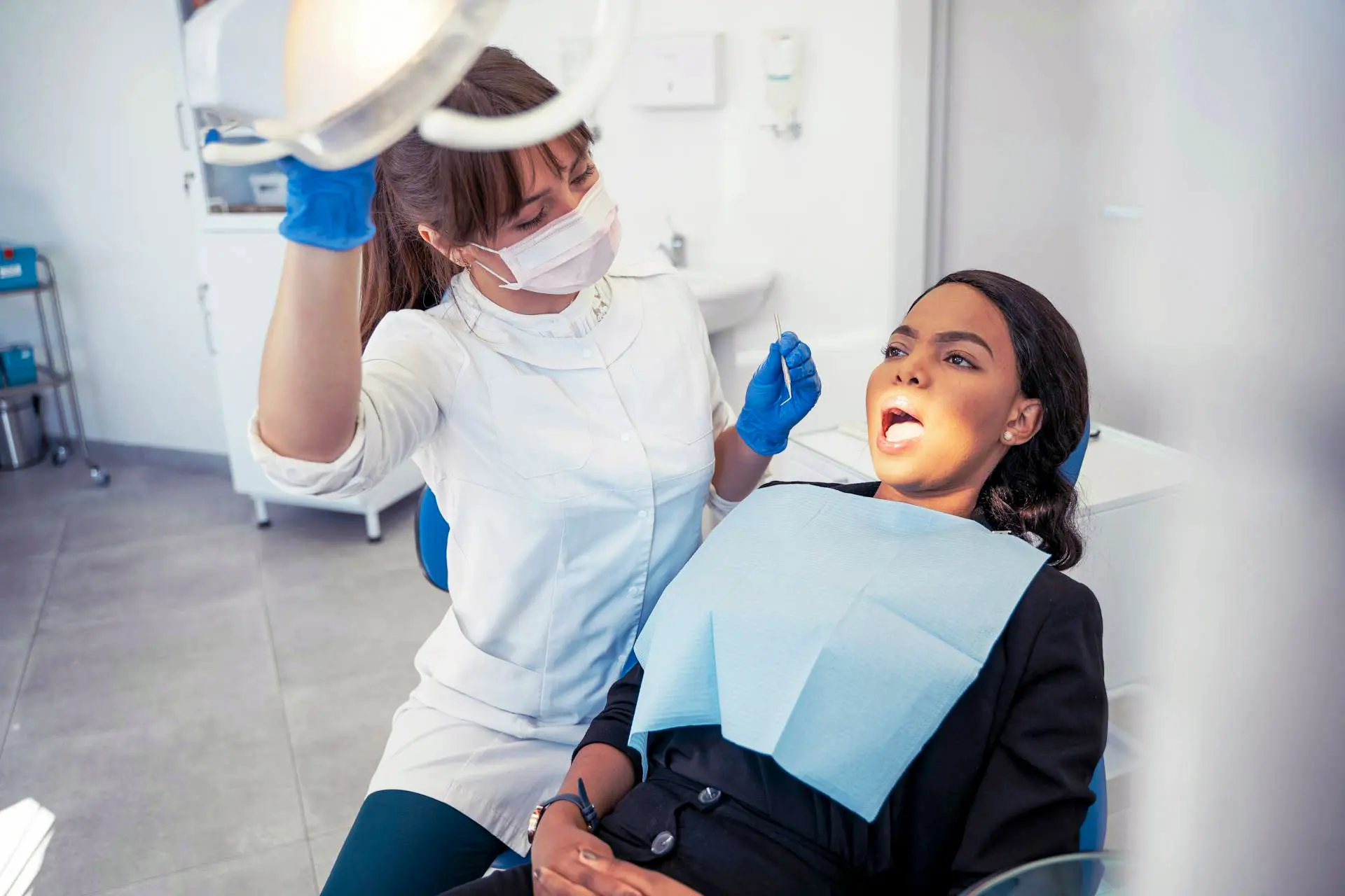 A dentist in gloves examines a patient lying back in a dental chair, mouth open, under bright overhead lights.
