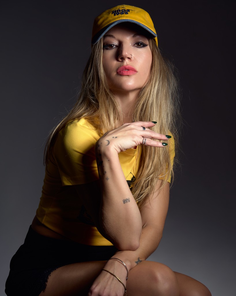 Una persona con una gorra y una camiseta amarillas posa con confianza, sentada con una mano en la barbilla frente a un fondo oscuro