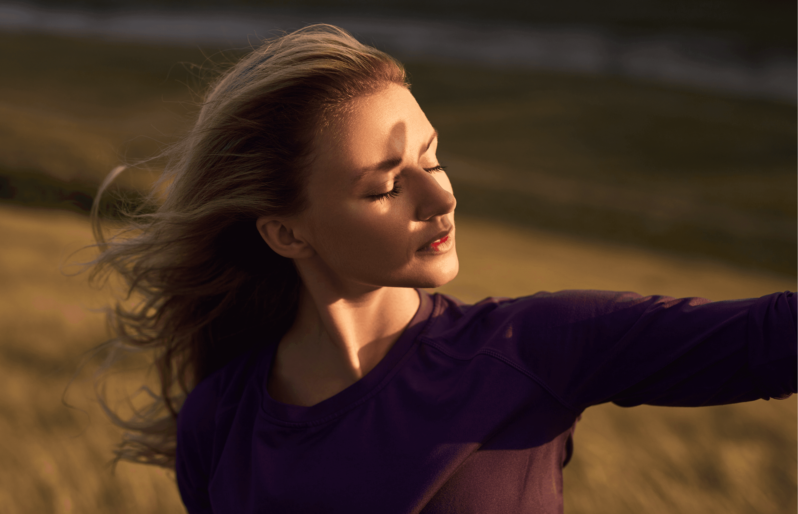 A woman with flowing hair, eyes closed, basks in warm sunset light in a serene field. She wears a purple top, exuding tranquility and contentment.