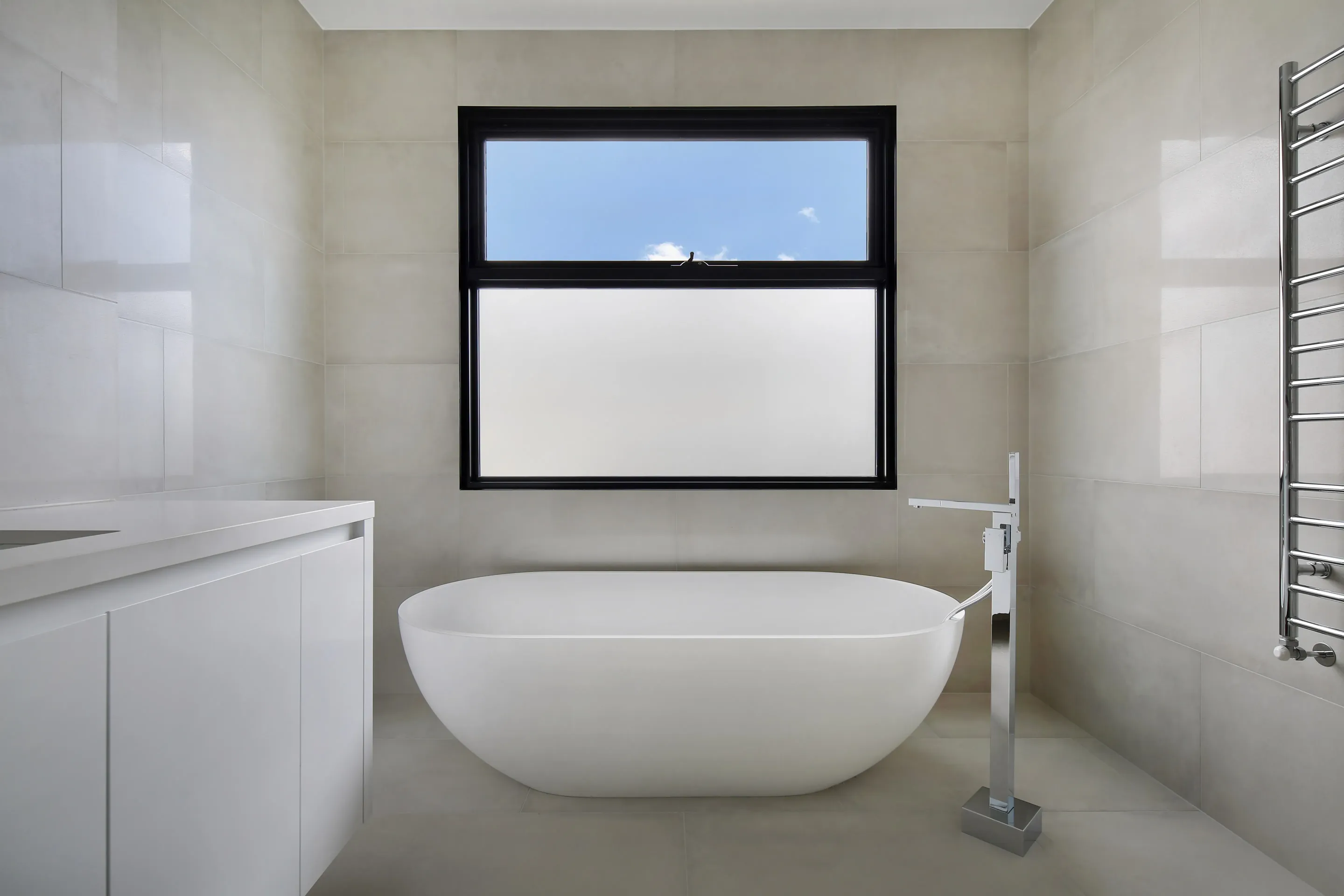 Bathroom in beige stone-look tiles with a white freestanding oval bath, a floor-mounted chrome bath filler, white vanity, chrome towel rail and a black-framed window with frosted lower pane