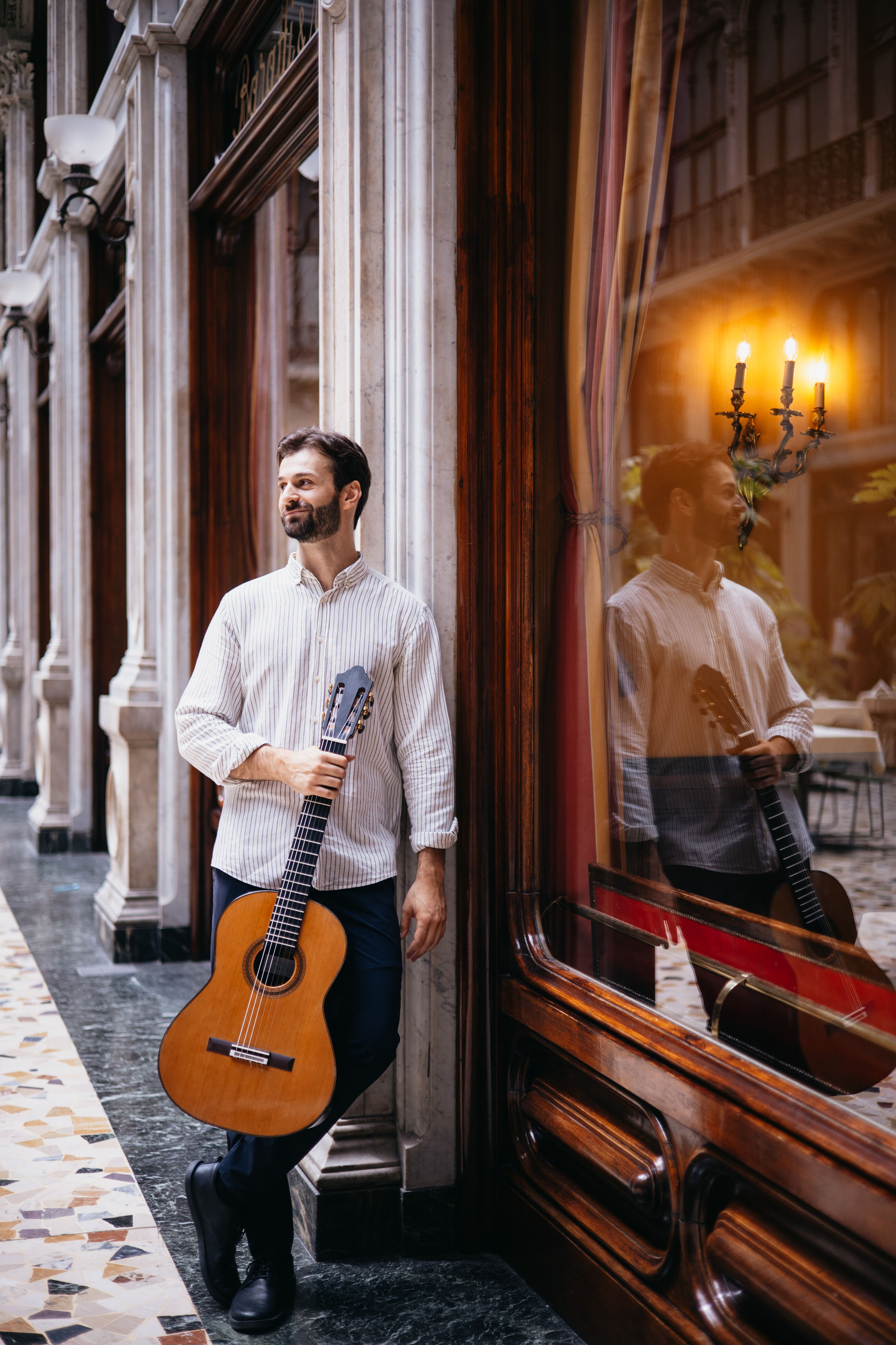 Andrea Curiale with his guitar beside a window, reflected in the glass