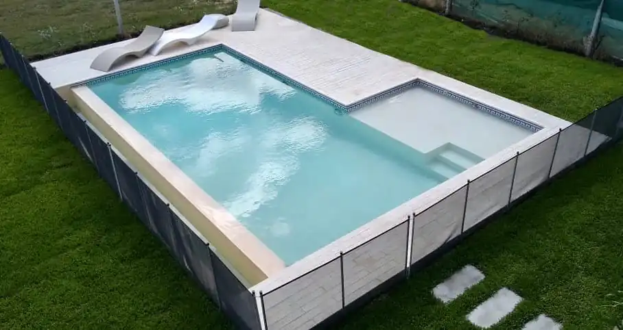 Pileta de diseño con solárium blanco, reposeras minimalistas y un prolijo cerco perimetral de seguridad oscuro en un jardín de césped.