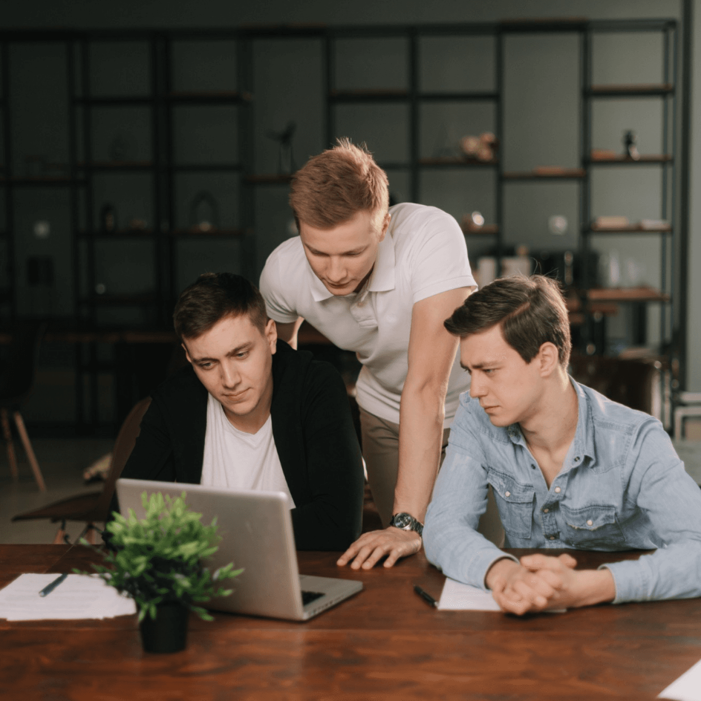 Three people collaborating around a laptop at a table with papers and a plant