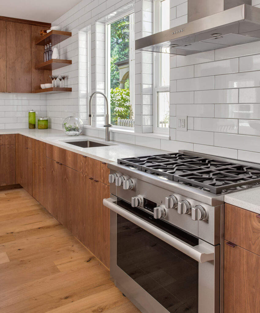 Modern kitchen with white walls and backsplash, natural wood cabinets, a white island with a waterfall countertop, and four metal barstools. Two round pendant lights hang above the island, and a stainless steel oven and hood are installed along the back wall with windows above the countertop.