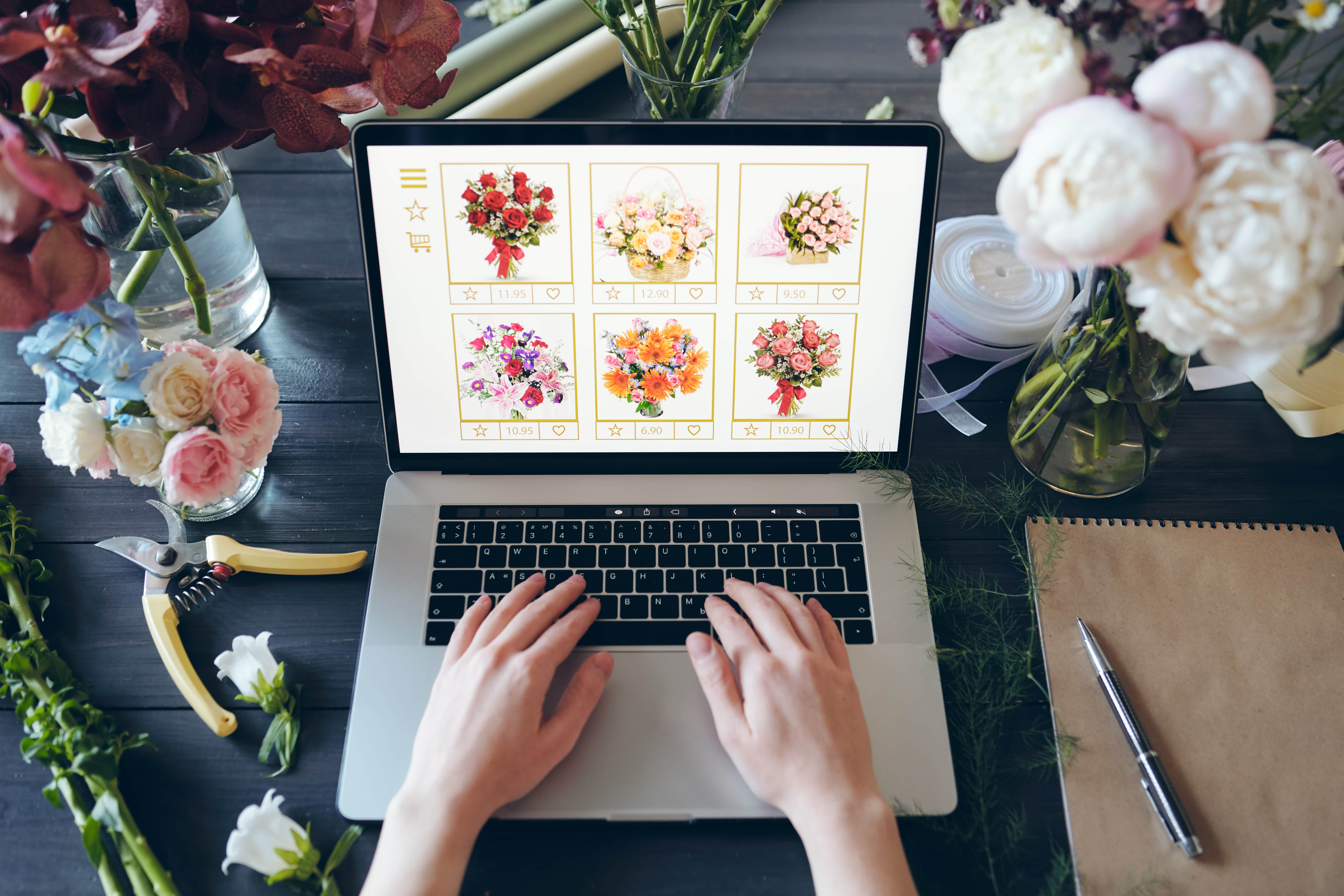 A top-down view of a florist working on a laptop, managing their online flower shop. The screen displays an e-commerce website with various bouquets for sale. The dark wooden desk is surrounded by fresh flower arrangements, including peonies and roses, along with florist tools like pruning shears and a notepad, showcasing a modern small business.