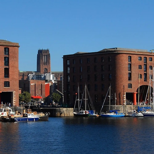 맑은 파란 하늘 아래 물가에 정박한 돛단배와 붉은 벽돌 건물들. 배경에는 높이 솟은 교회탑이 보입니다.