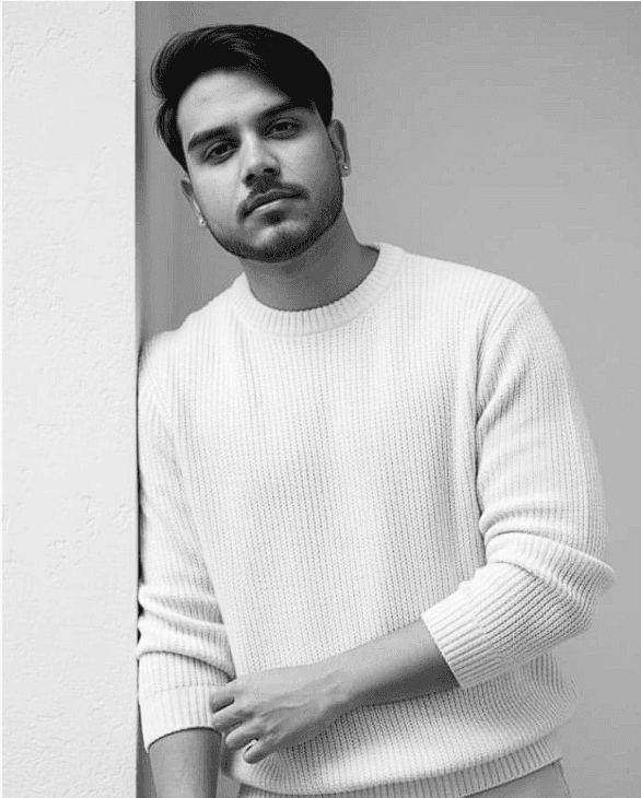 A young woman with long hair smiles while standing against a green backdrop, wearing a casual dark sweater.