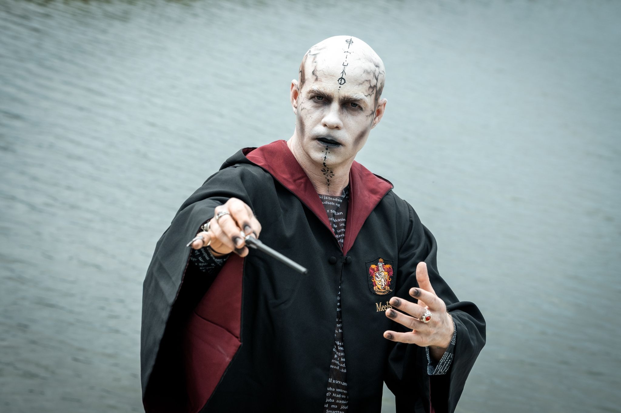 Thom Rikko in theatrical makeup and a dark robe, holding a wand toward the camera.