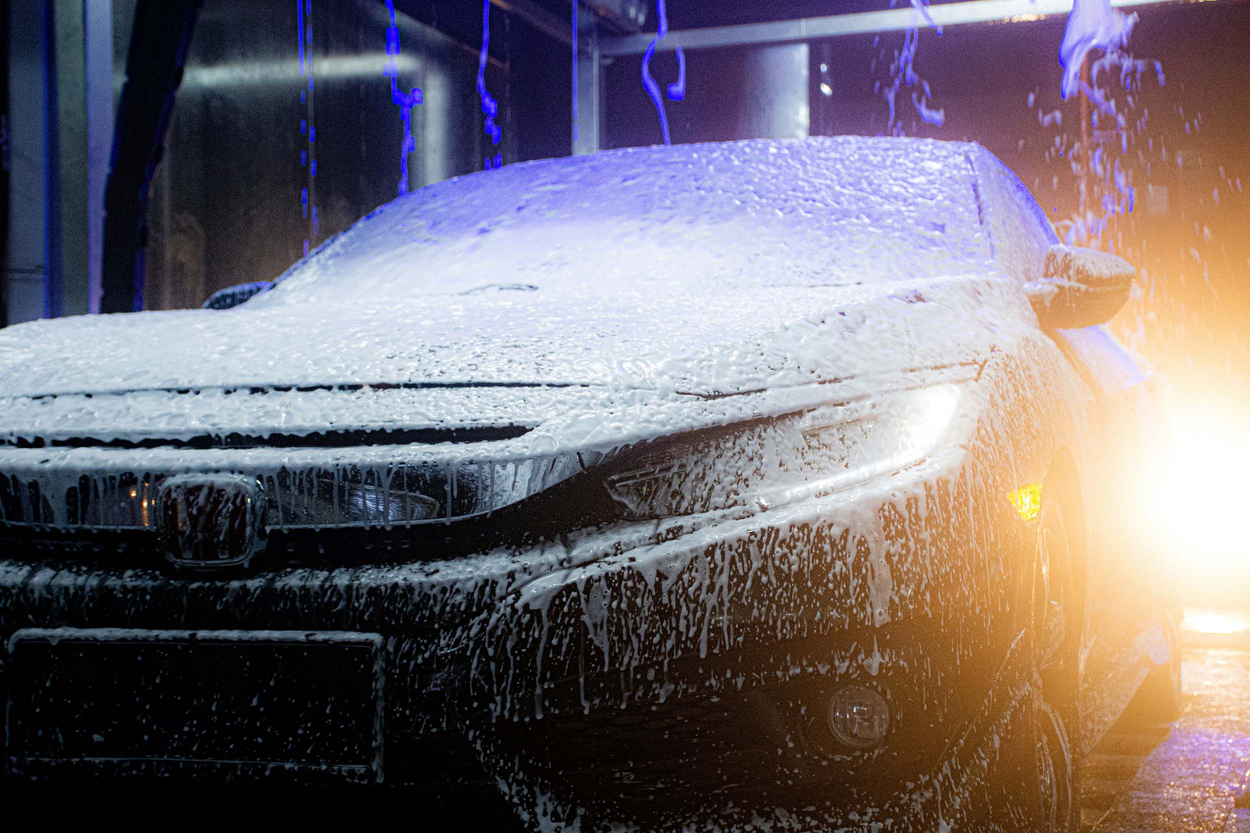 A close-up of a black Honda car being washed in an automated car wash. The car's front end is covered in thick white foam, with water droplets streaming down the hood and bumper. The headlights are illuminated, casting a warm glow through the foam, while blue and white lights from the car wash equipment create a dramatic effect in the background. The scene is dynamic, with water and foam in motion, highlighting the cleaning process.