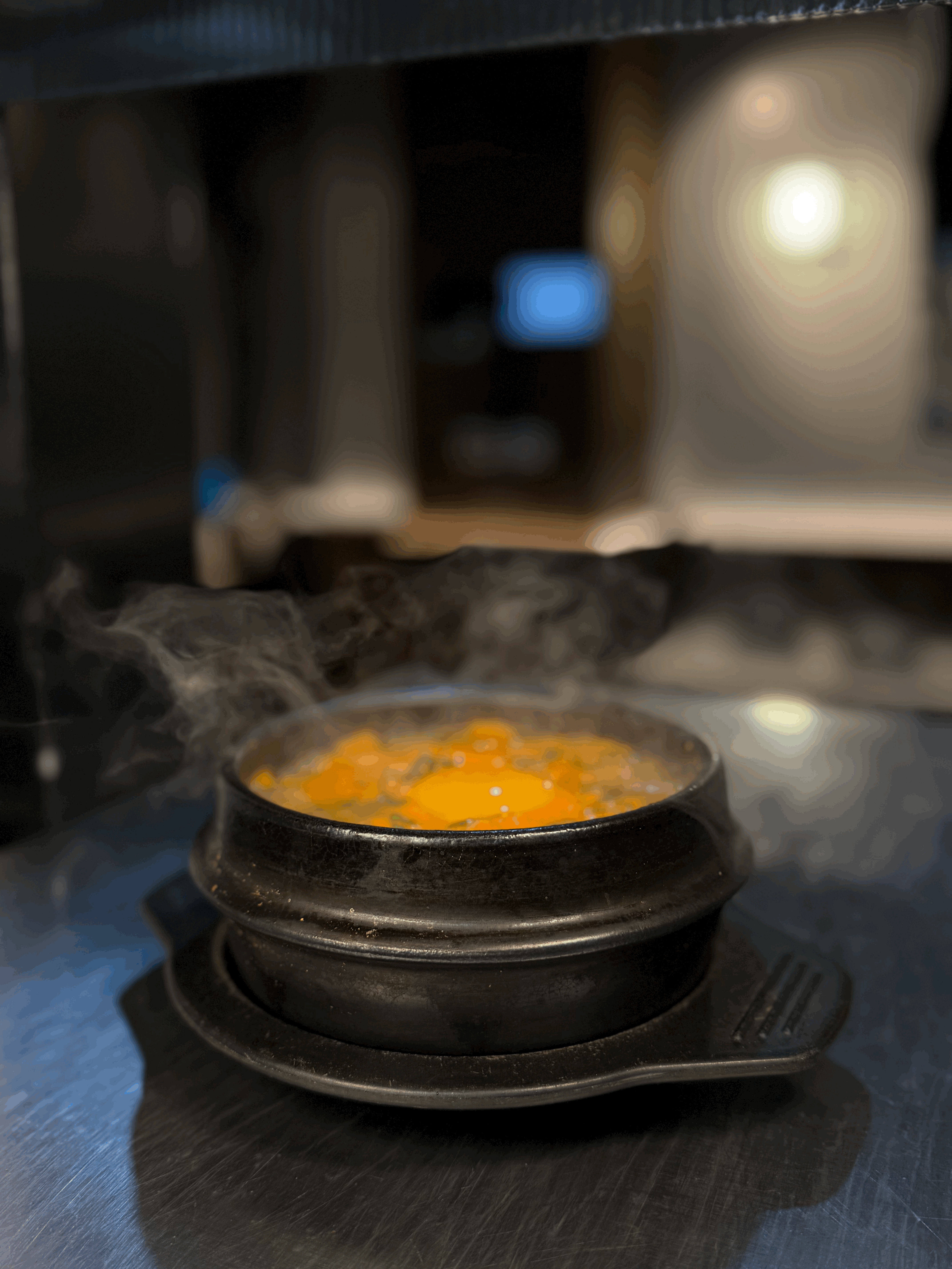 A close-up of a dark gray bowl filled with white fish chunks, cherry tomatoes, green olives, and herbs, served next to a glass of white wine on a white table by a window.
