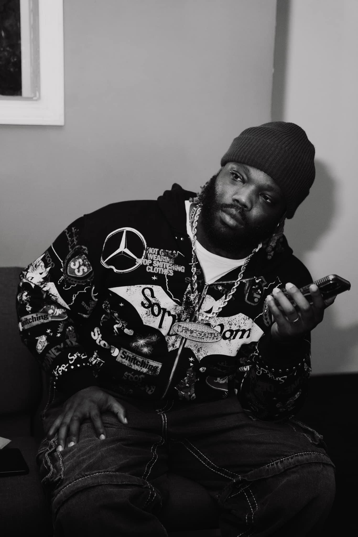  HitKidd, a renowned Memphis producer, sits contemplatively holding a smartphone. Dressed in a patch-adorned jacket and knit cap, this black-and-white image captures him in a moment of reflection, possibly during an interview where he discusses his career and creative process.