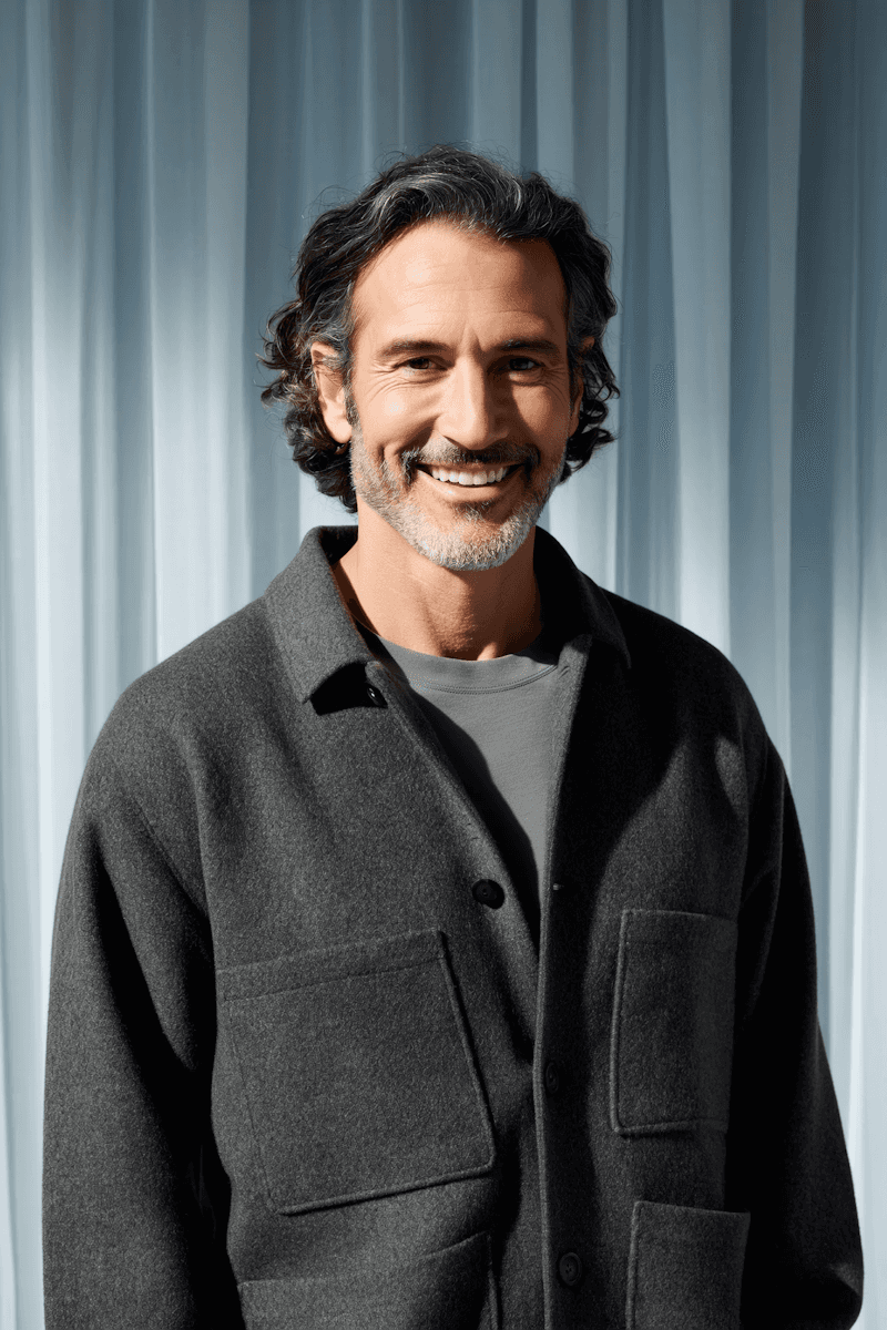 A smiling man with dark hair, wearing a black shirt, poses against a soft blue background.