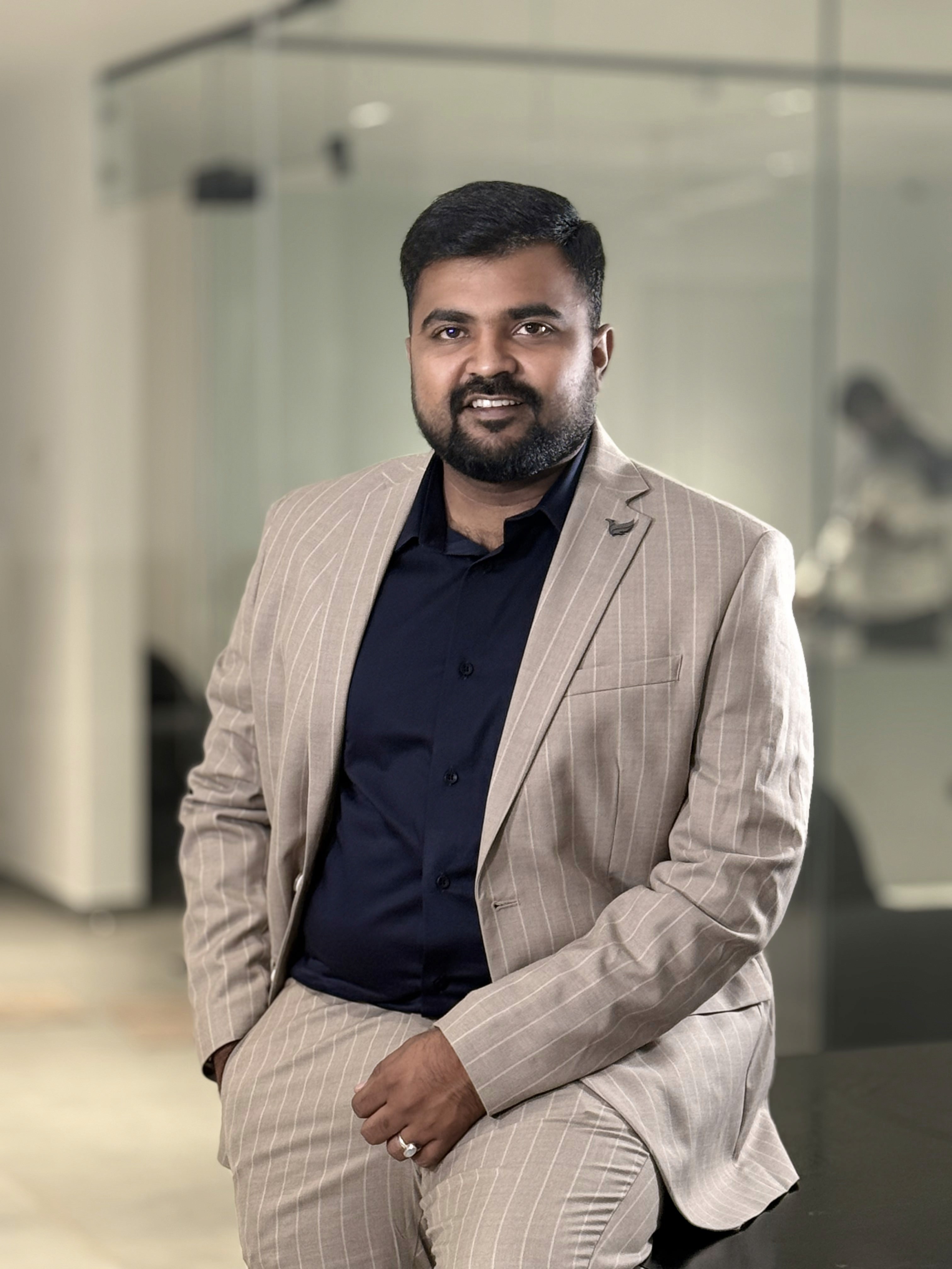 Young man in a dark suit jacket and cream sweater vest over a blue shirt with a patterned tie. Professional and confident expression, neutral background.