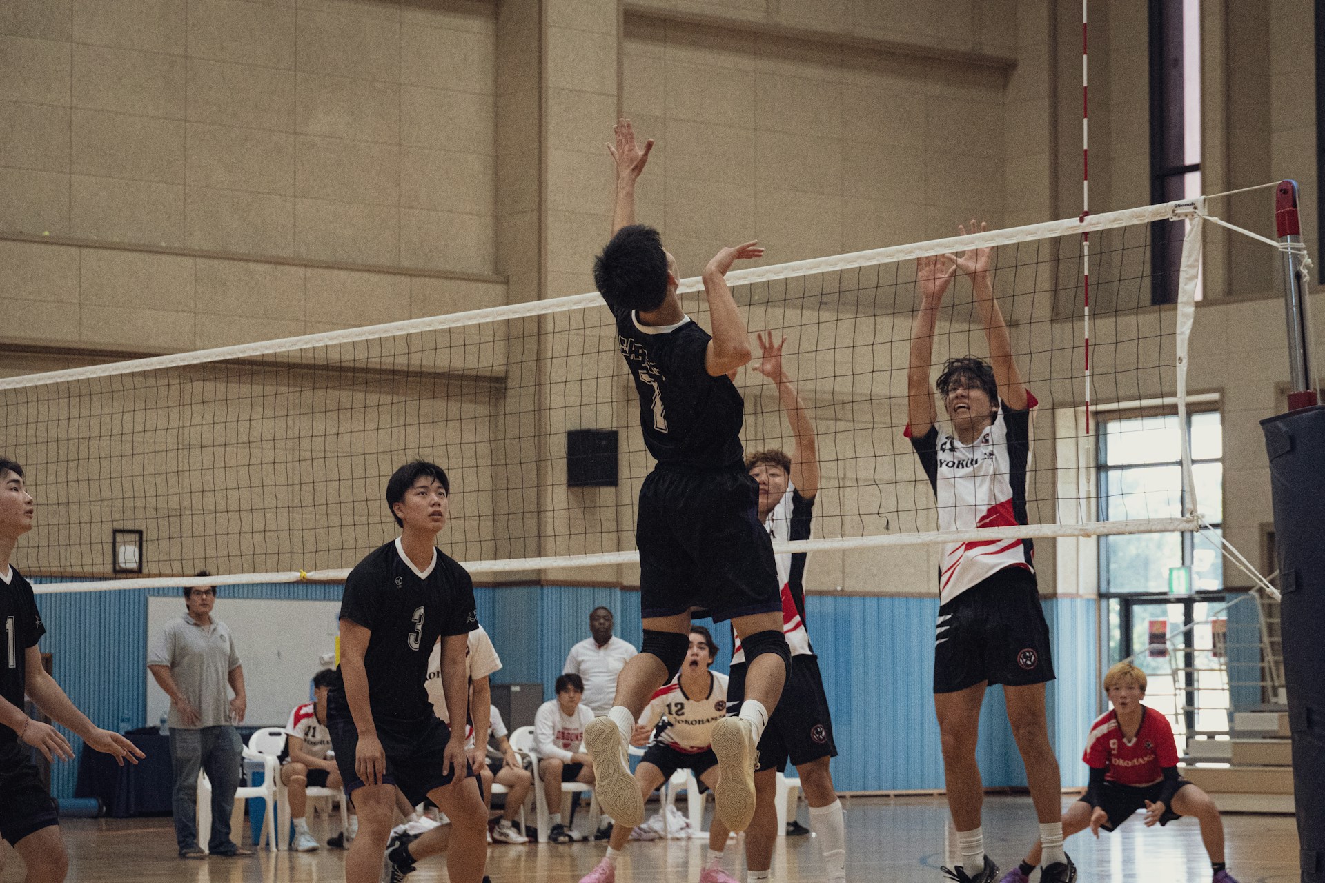 A volleyball player in a black uniform jumping high to spike the ball at the net while opponents attempt to block.