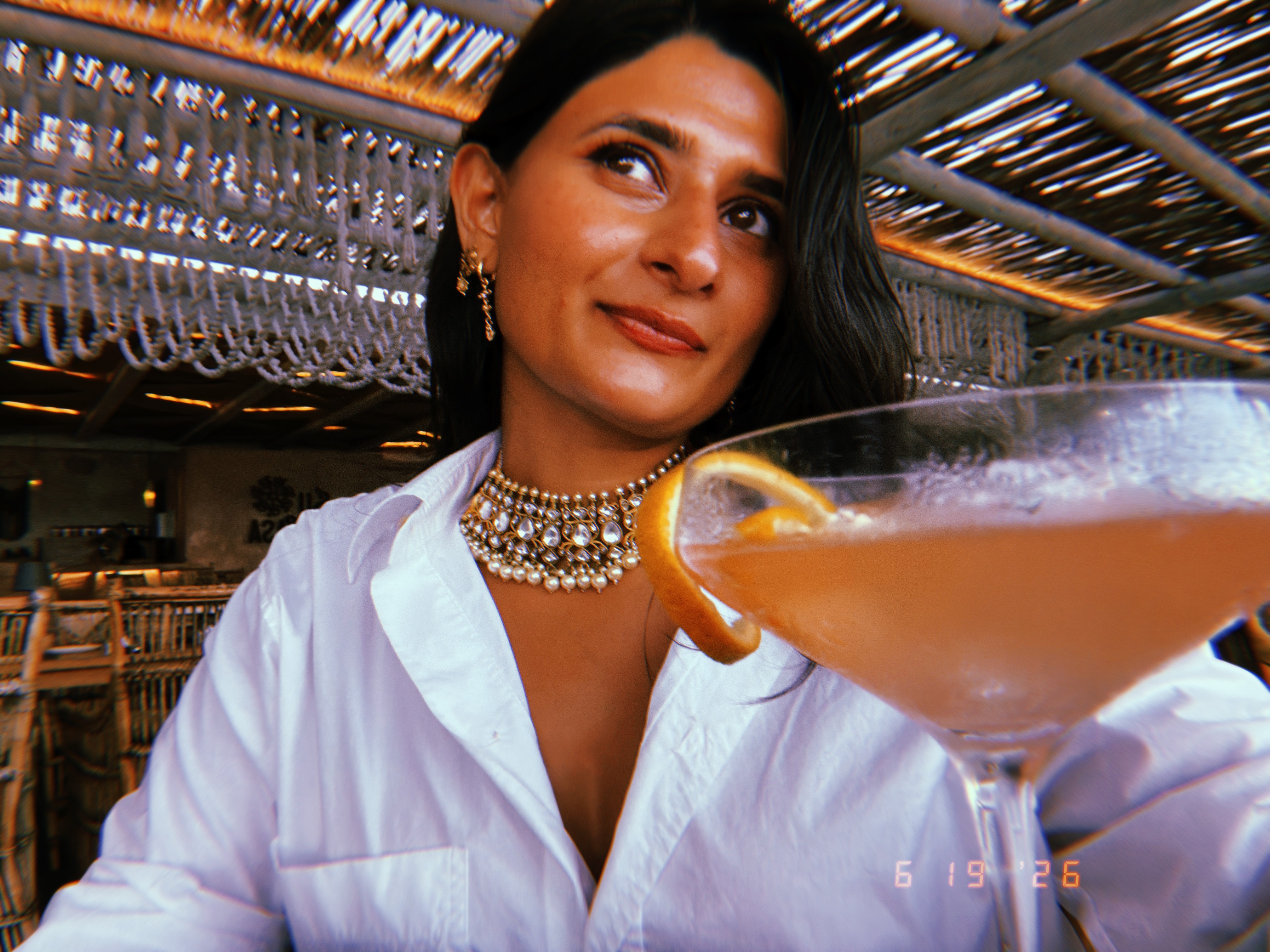 Pranjali in a white shirt and jeweled necklace, with a coupe glass in the foreground
