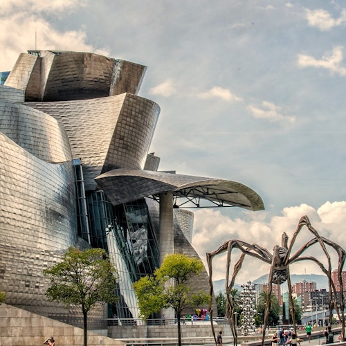 Modern, metallic building with curved architecture. Large spider sculpture and people walking nearby. Trees and a cloudy sky in the background.
