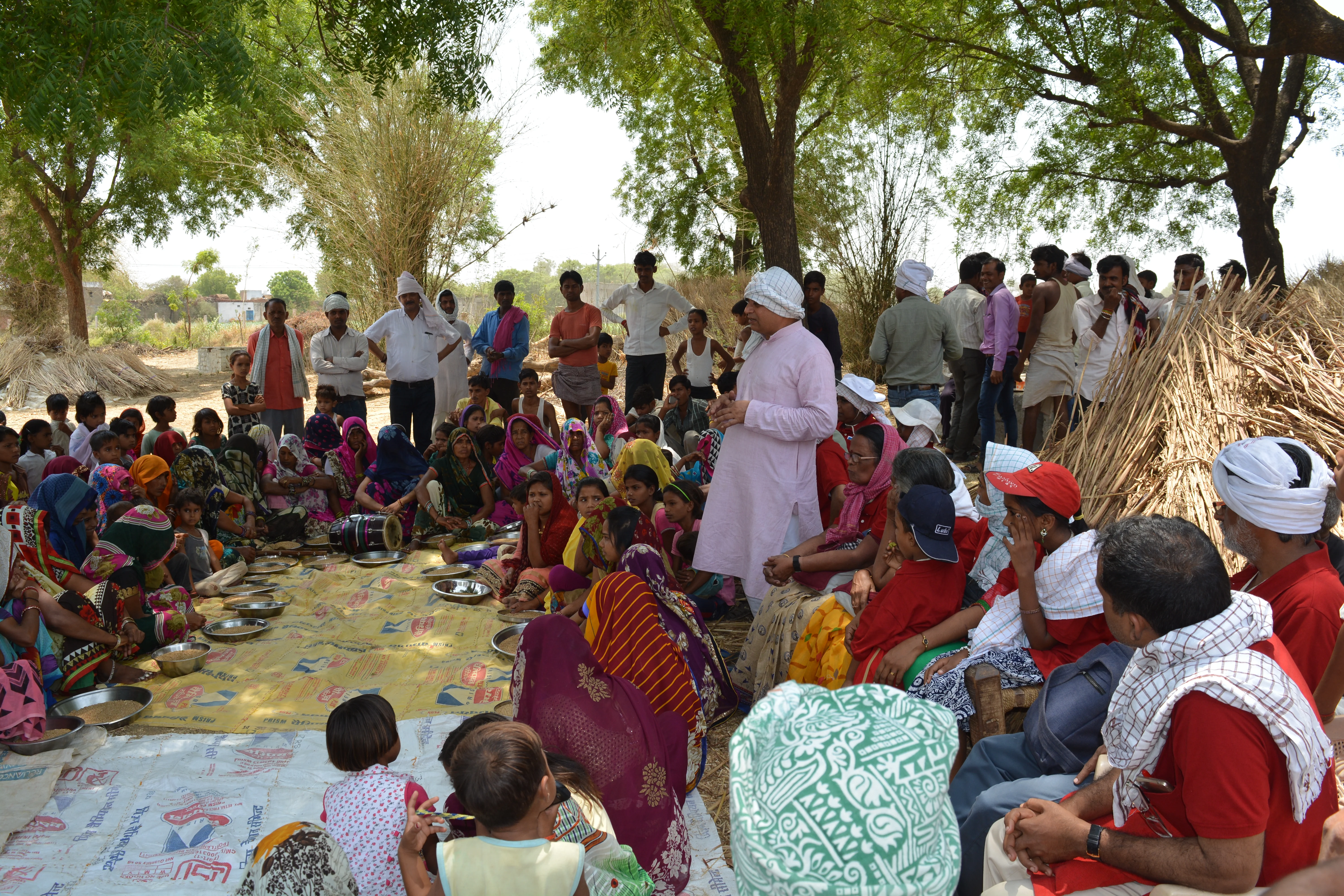 A large group of villagers, mostly women and children, gathered under trees in a rural setting, sitting in a circle on mats for a meeting or discussion.