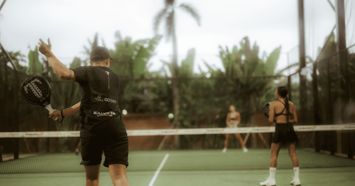 Players in a doubles match on a green padel court surrounded by tropical plants and palm trees.