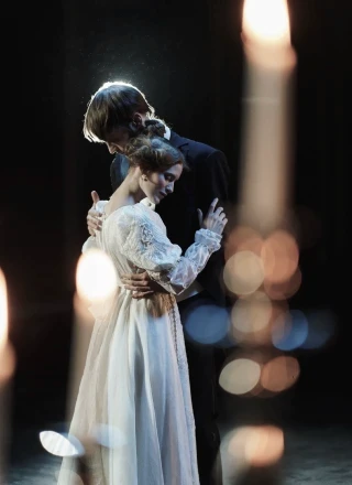 Two dancers in period costumes embrace on a dark stage