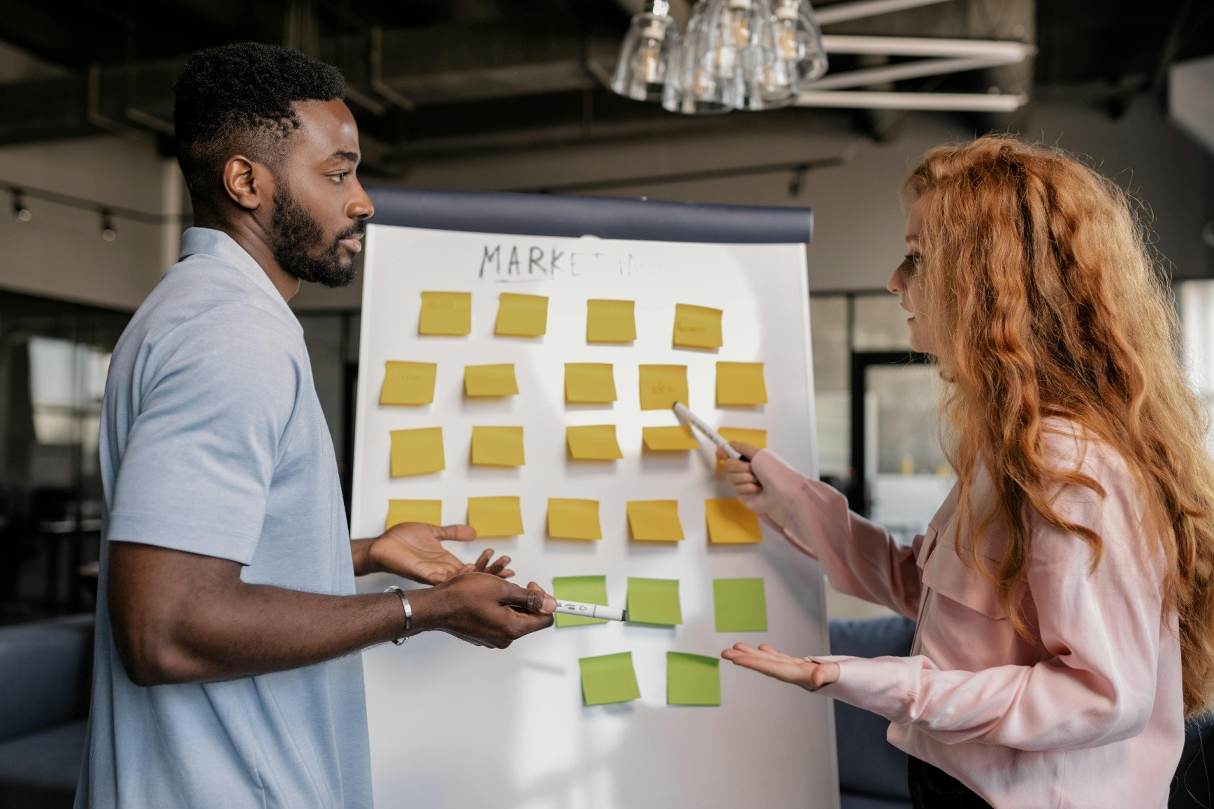 Two colleagues collaborate in a modern office, discussing ideas on a whiteboard filled with yellow and green sticky notes. The woman gestures to a note with a marker, while the man listens attentively, marker in hand