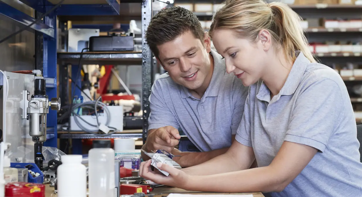 Zwei Personen arbeiten gemeinsam an einem Arbeitsplatz und besprechen ein Projekt, während sie ein Gerät oder Werkzeug untersuchen.