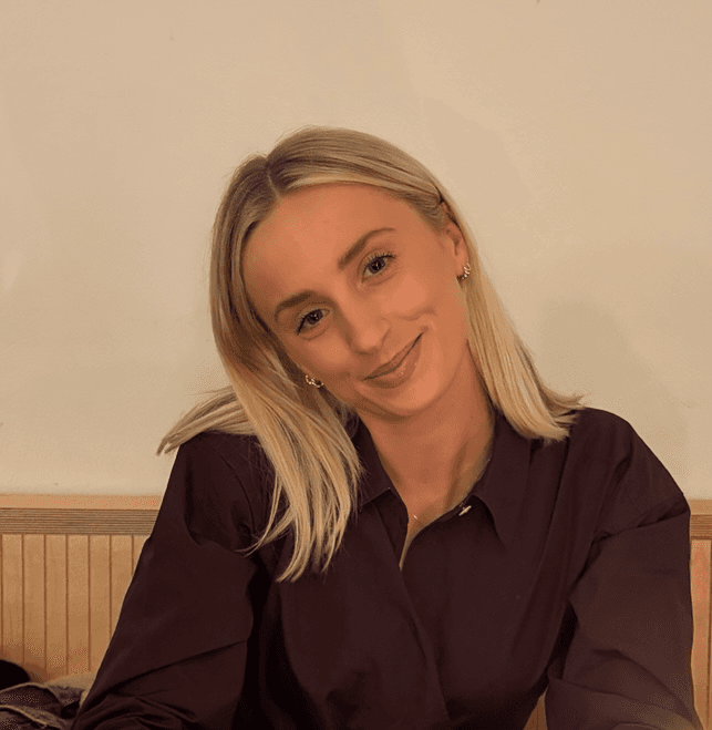 Portrait of Alexis Hedley, Campaign Manager at Playkit, smiling in front of a beige wall and wearing a black strapless top with a gold cross necklace.