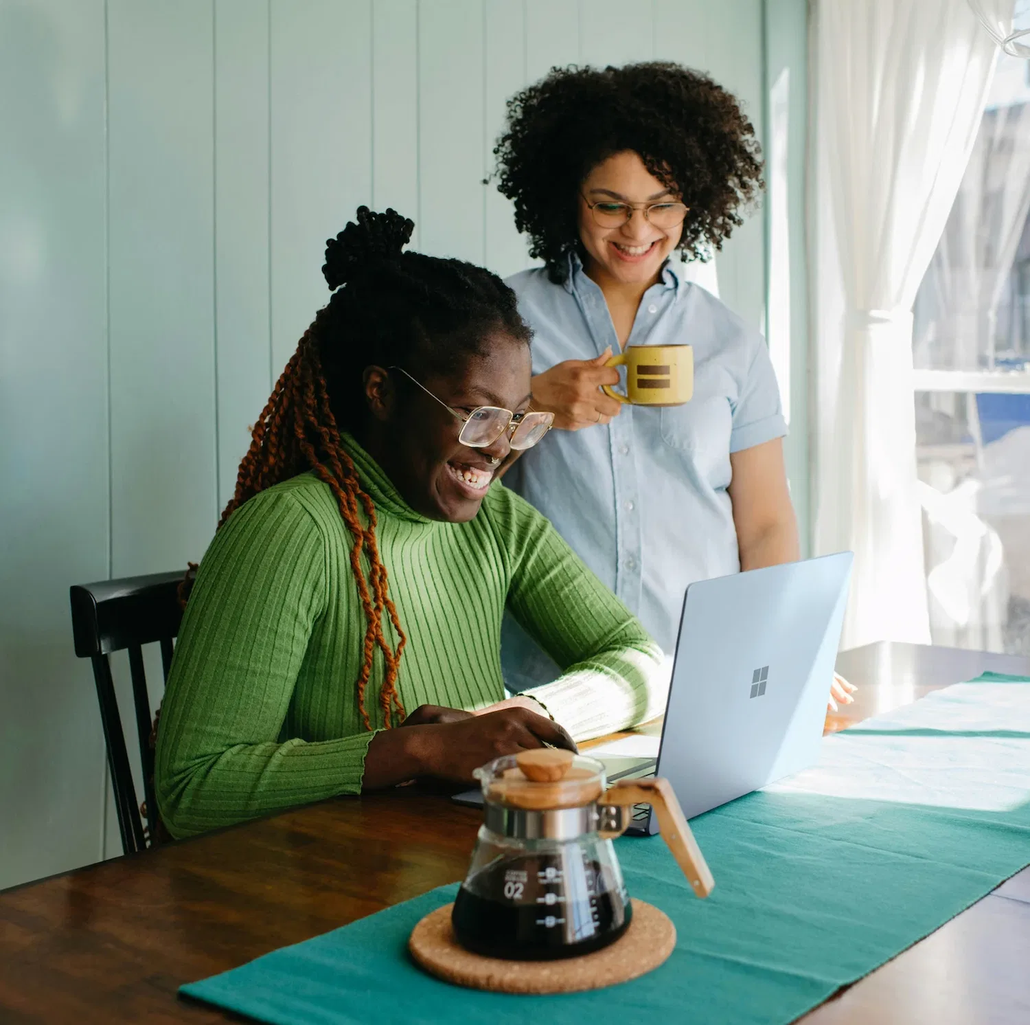 Two women enjoying coffee and smiling together while using a laptop during an online therapy session at home in New York City.