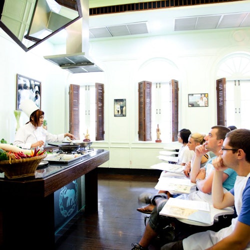 Seorang chef sedang menunjukkan teknik memasak kepada sekumpulan pelajar yang tekun di dalam kelas dapur yang terang benderang.