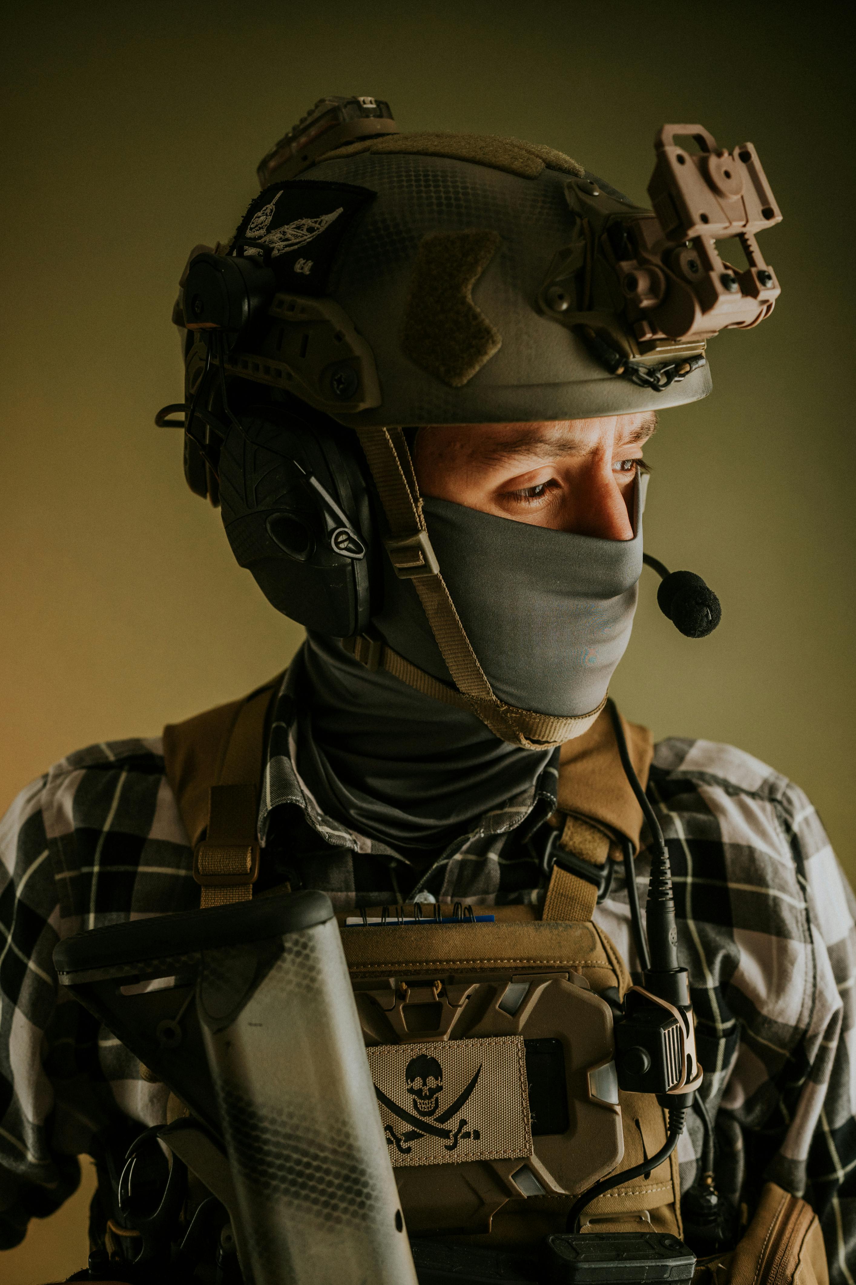A soldier wearing a tactical helmet with night vision equipment mounted on top. The person is wearing a camouflage uniform with a face mask and tactical vest with various pouches and equipment including visible orange scissors. The image shows military personnel in full combat gear.