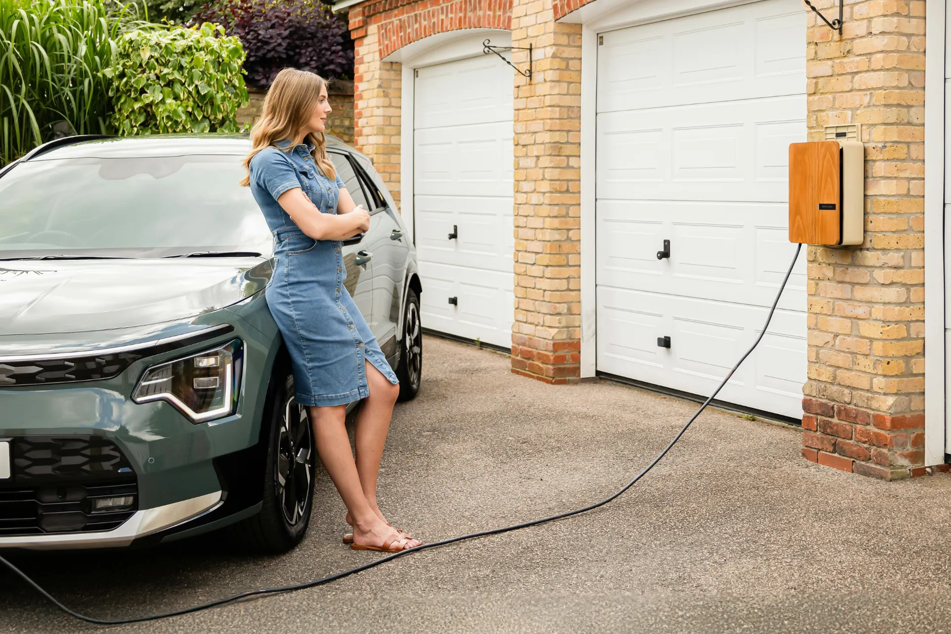 Eine Frau die an angelehnt an ihrem Auto steht. Das Auto ist gerade an der Wallbox befestigt welche an der Garagenwand befestigt ist.
