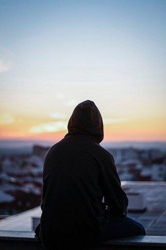 Ambigous photo of a man in a hoodie sitting on a building looking out over a city