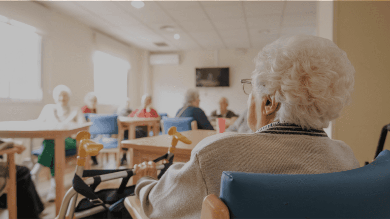 An elderly person seated in a care home communal room, viewed from behind, with other residents and tables visible in the background