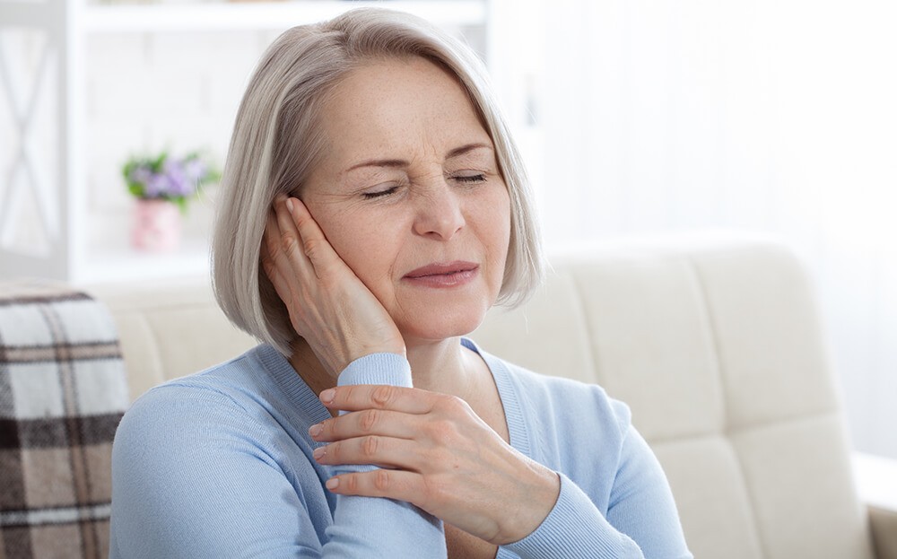 A woman with short gray hair sits on a couch, holding her cheek, appearing to be in discomfort.