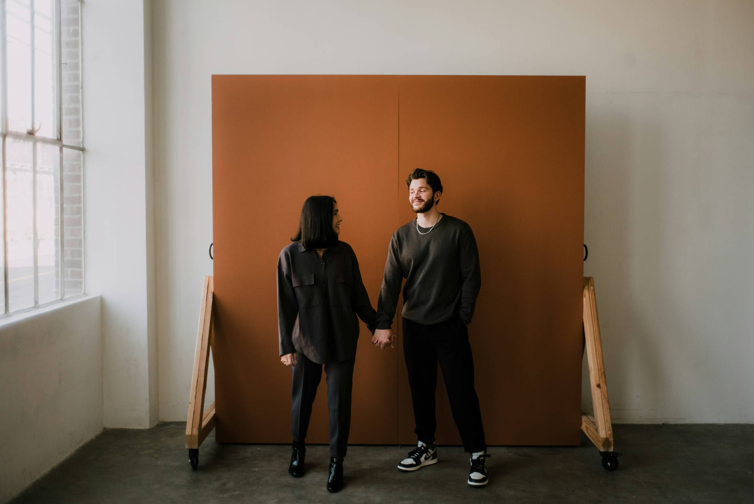 A couple holds hands in front of an orange studio backdrop with a warehouse style window to their right