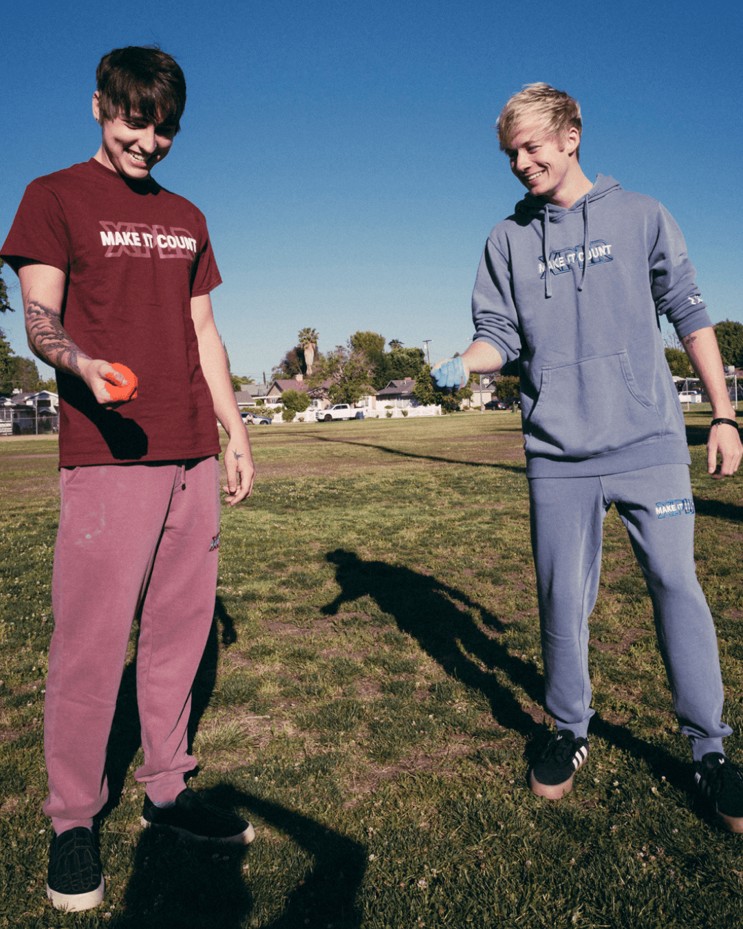Two young men stand on grass, holding red objects, smiling under a clear blue sky.