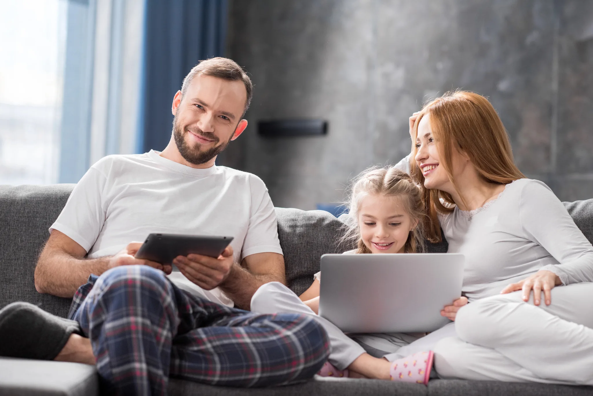 Happy family sitting on a couch, Husband and daughter holding a device, Wife looking at husband, Family wearing comfortable clothes.