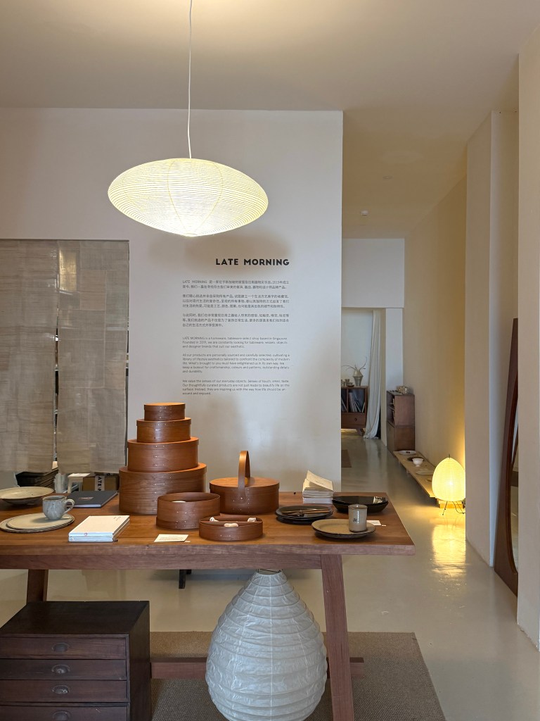 Interior of a minimalist shop named 'Late Morning' featuring a wooden table with ceramics, stacked wooden containers, and a soft pendant light.