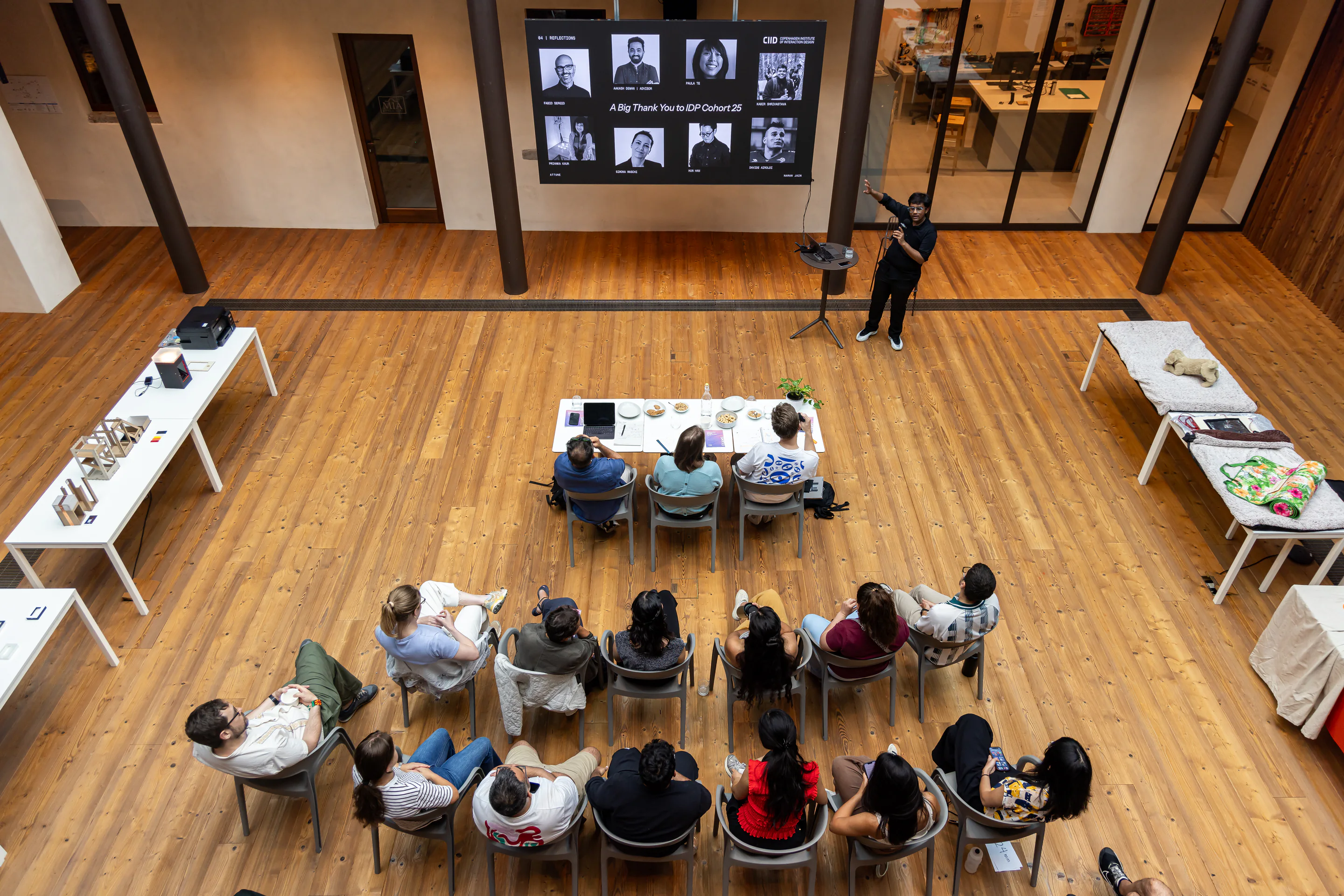 Overhead view of a presenter addressing a seated audience in a wood-floored atrium, with project work displayed on surrounding tables.