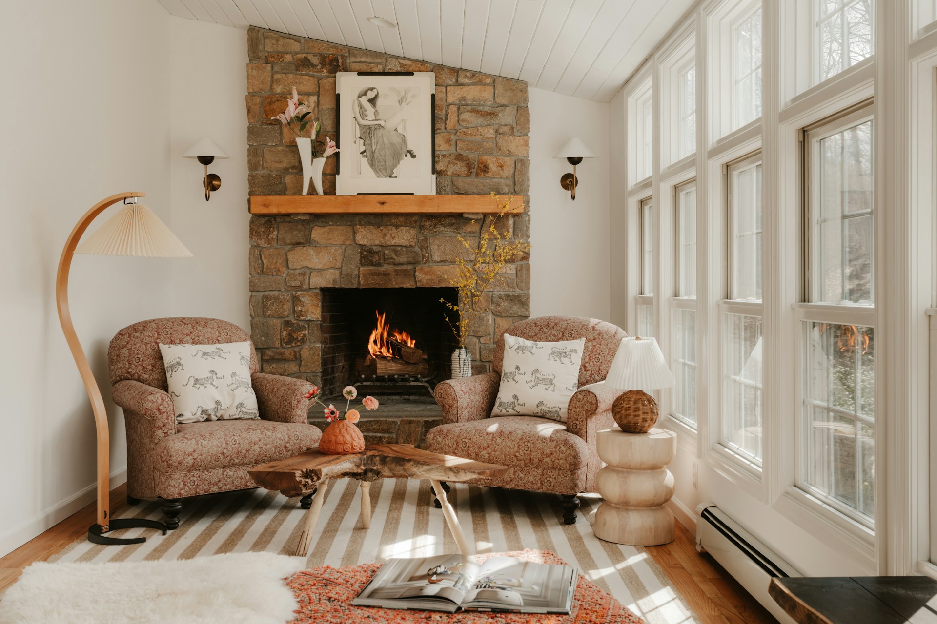 Cozy living room featuring a stone fireplace, two patterned armchairs, a live-edge coffee table, and large sunlit windows.