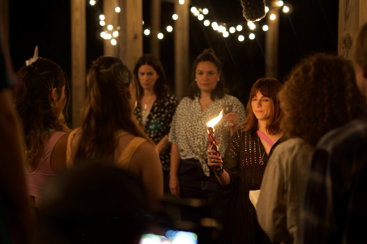 A group of women stands in a dimly lit outdoor setting, one holding a lit candle, with string lights hanging in the background.