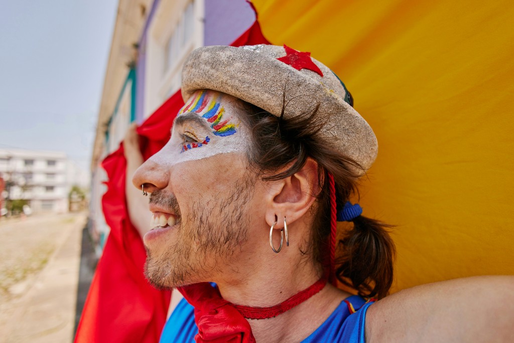 A cheerful performer with colorful face paint and a decorated hat smiles brightly, exuding joy and creativity during a vibrant street festival.