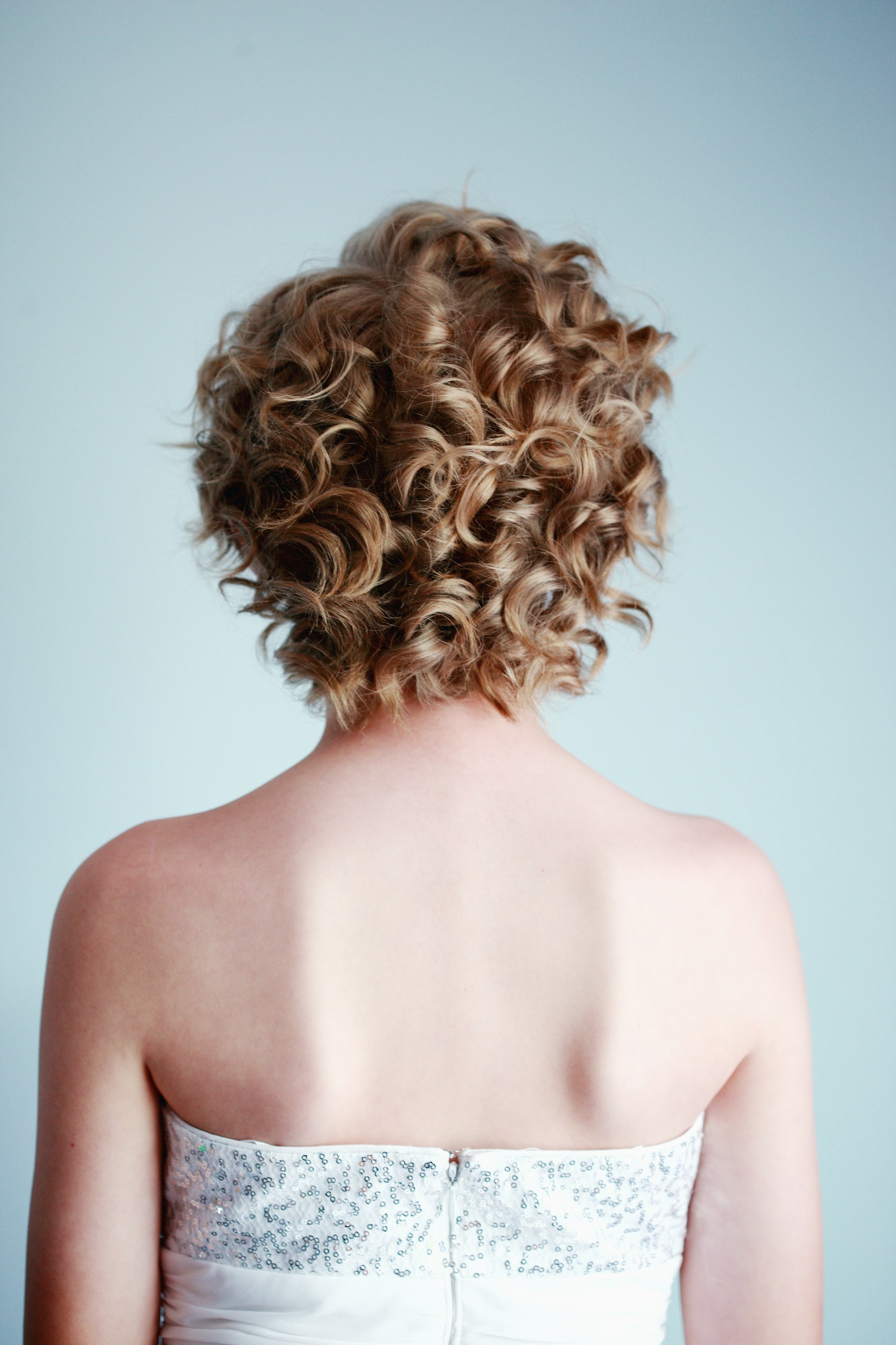 The back of a person with curly, medium-length blonde hair, wearing a white, strapless, sequined top against a solid light blue background.