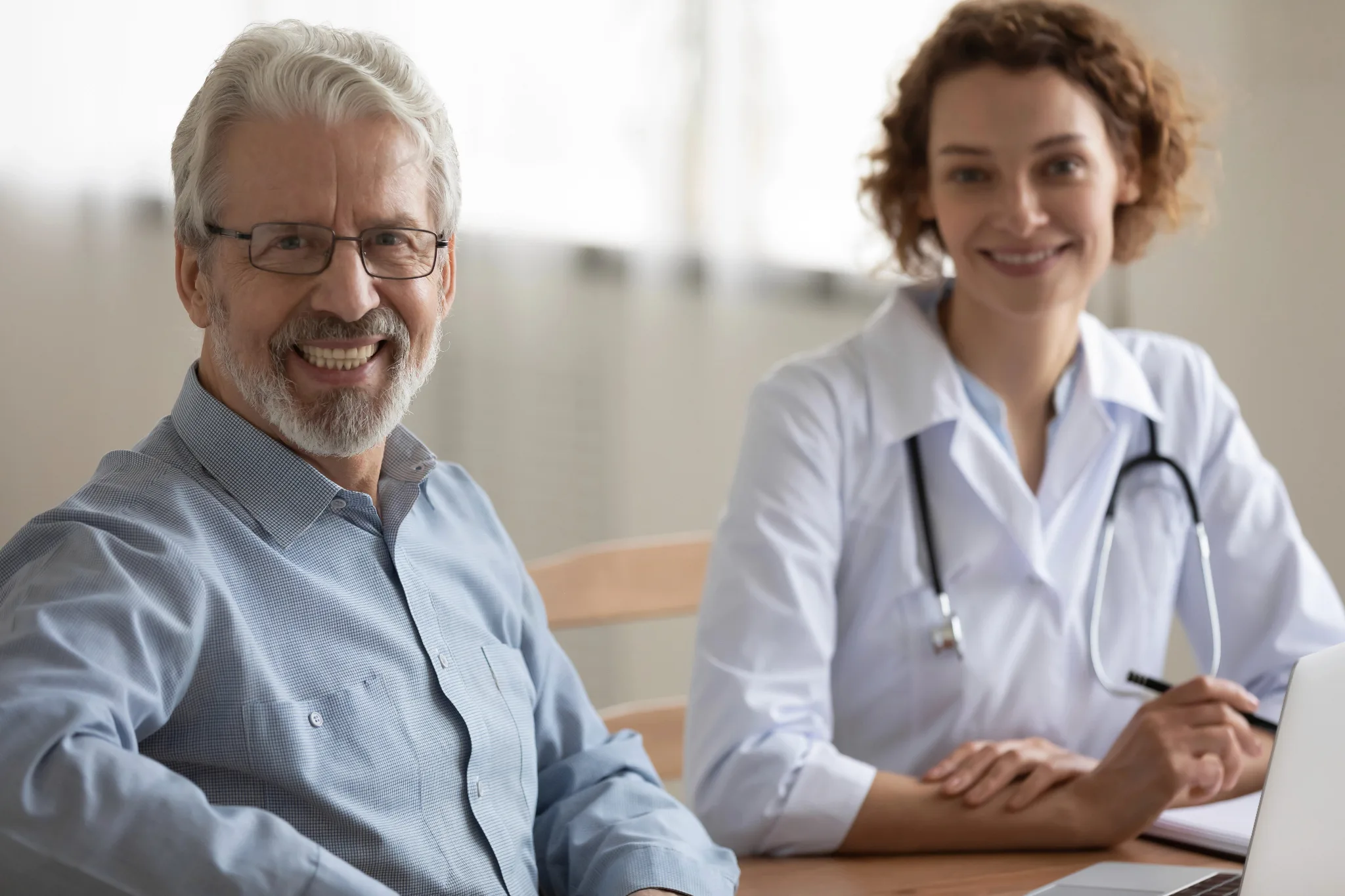 Smiling female doctor wearing a white coat with stethoscope around neck, sitting on a chair with two arms resting on a table, a male patient with white hair and glasses wearing a formal attire sitting beside the physician.