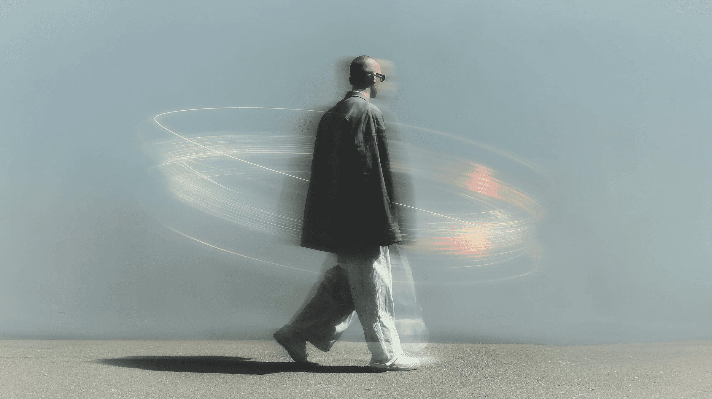 Bald man in grey coat walking against a light grey wall, slow shutter speed creating orbital light trail around him