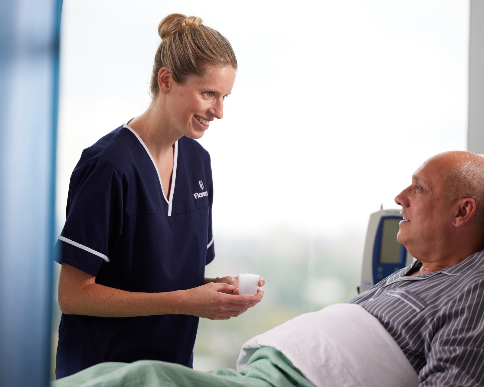 Nurse in navy Florence scrubs smiling while offering a small cup to a patient sitting up in a hospital bed.