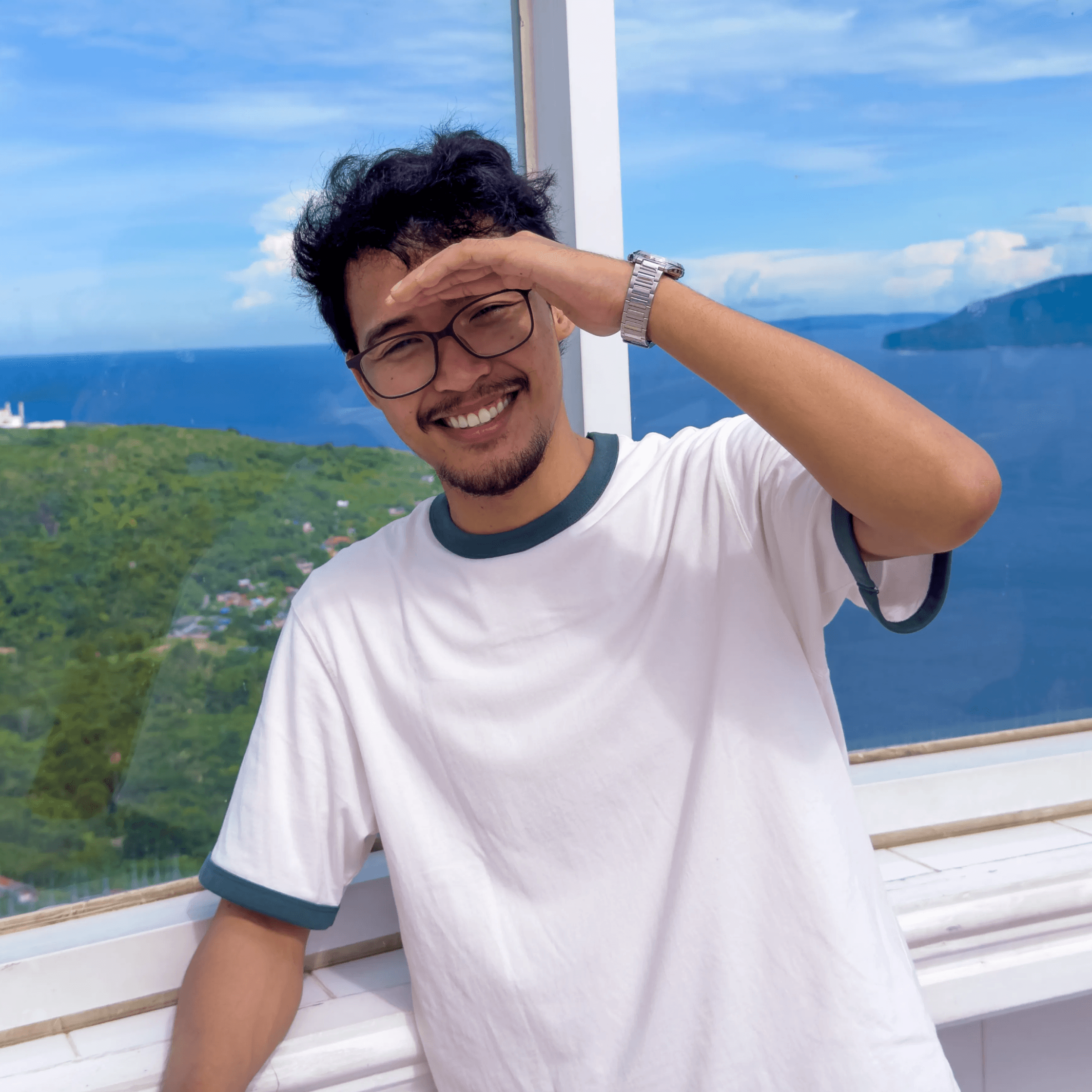 A man in glasses and a white t-shirt smiling and shielding his eyes with his hand near a scenic ocean view.