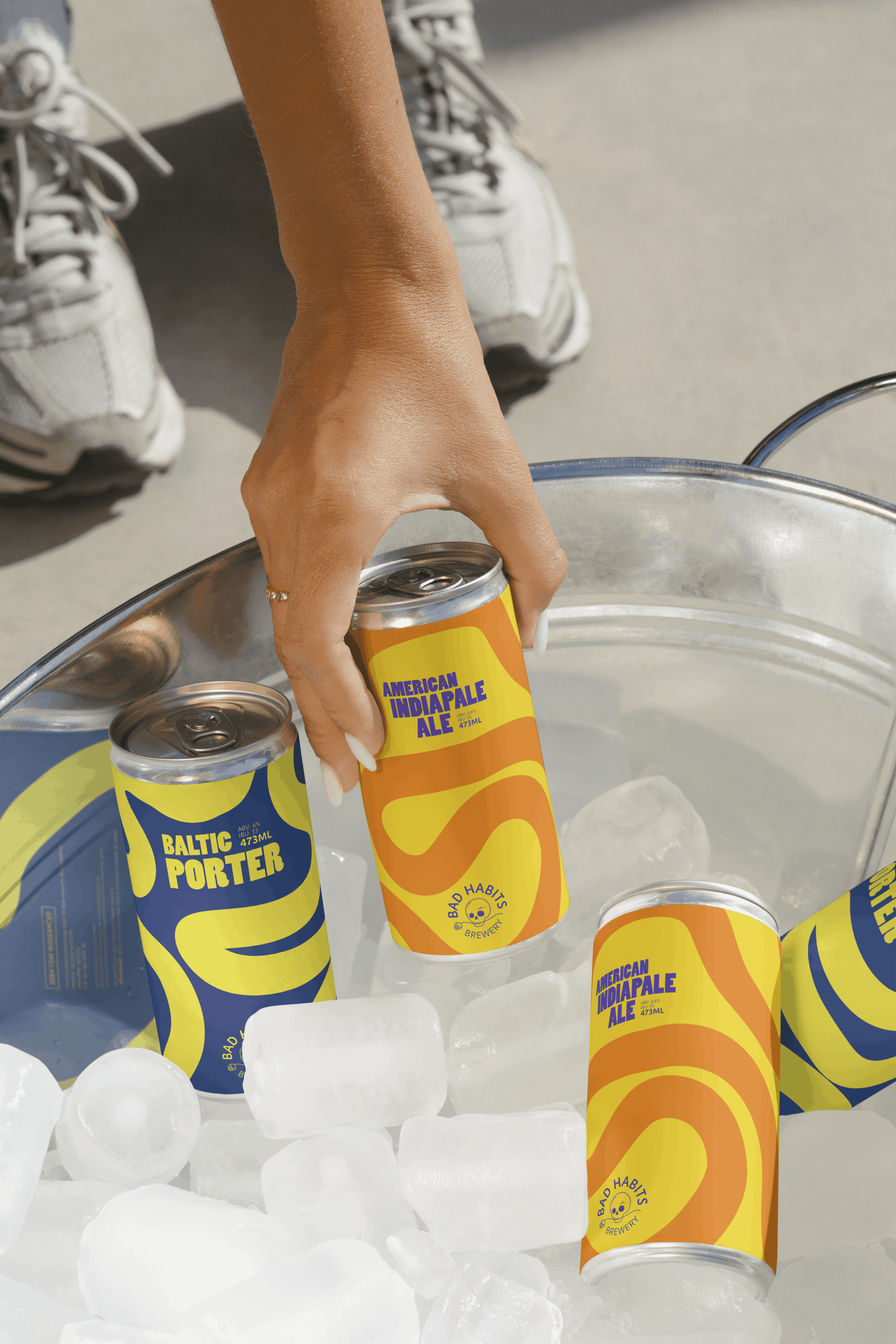 A hand lifting a can of Bad Habits Brewery American India Pale Ale out of a metal bucket filled with ice cubes, alongside cans of Baltic Porter.